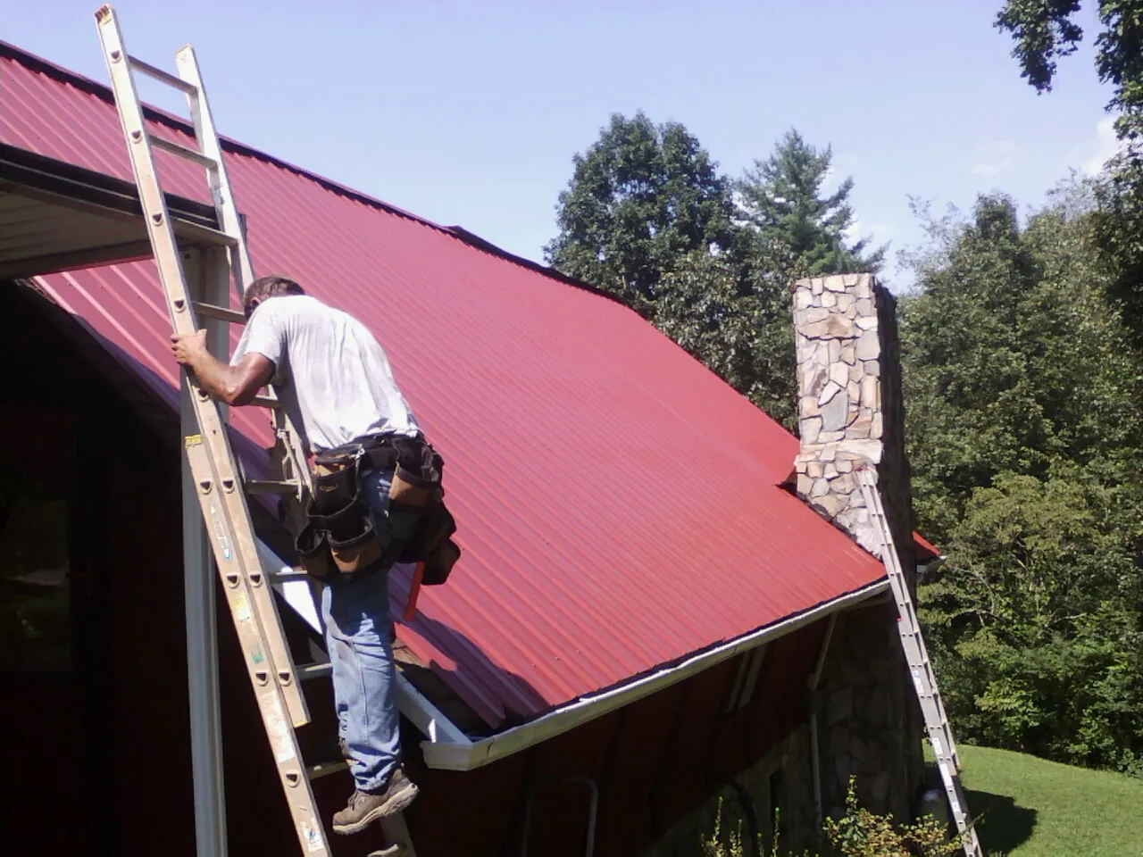 Roofer on a ladder beside a red metal roof and stone chimney — Soffit & Fascia Repair by Roofing Brothers in Hollow Rock